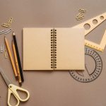 Caderno espiral, tesoura e outros materiais escolares organizados sobre uma superfície marrom.
