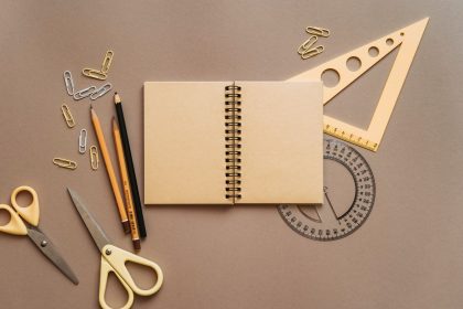 Caderno espiral, tesoura e outros materiais escolares organizados sobre uma superfície marrom.