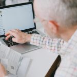 Idoso consulta documentos e administra suas finanças diante de um laptop em casa.