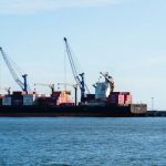 Navio cargueiro carregado de contêineres atracado em um porto industrial com guindastes.