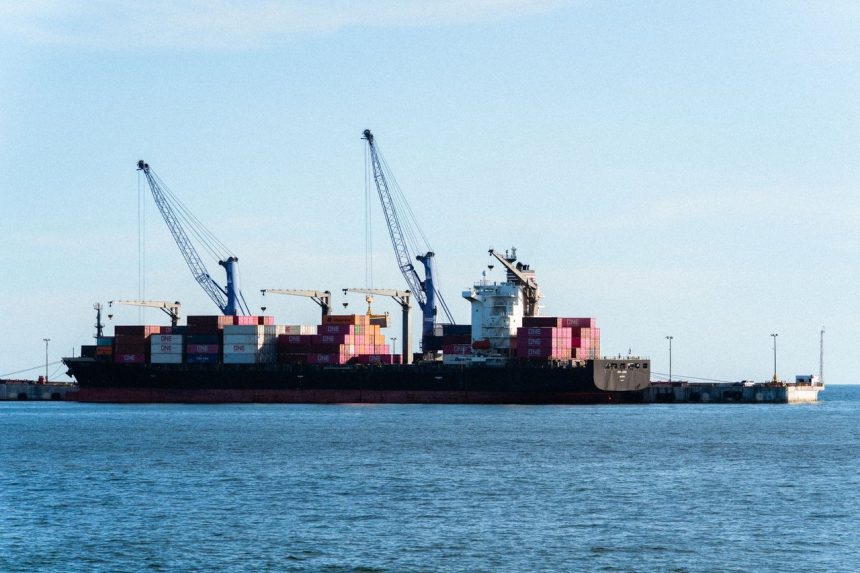 Navio cargueiro carregado de contêineres atracado em um porto industrial com guindastes.