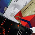 Cartões de crédito ao lado de um smartphone que exibe um gráfico do mercado financeiro.