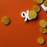 Moedas douradas sobre um gráfico do mercado financeiro, acompanhadas de um símbolo de porcentagem em fundo laranja.