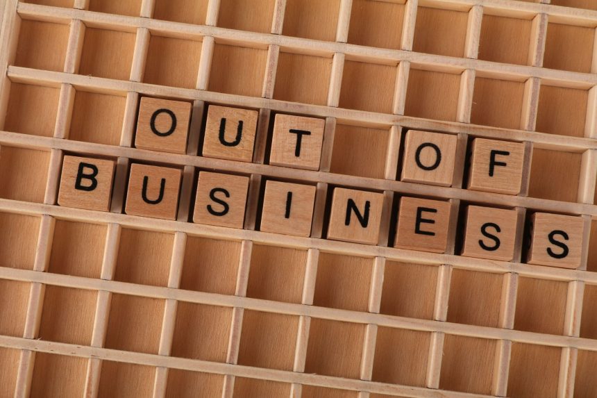 Blocos de madeira formam a expressão “Out of Business” sobre uma superfície de madeira, simbolizando o encerramento de uma atividade.