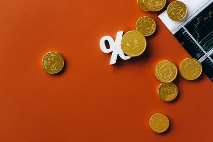 Moedas douradas sobre um gráfico do mercado financeiro, acompanhadas de um símbolo de porcentagem em fundo laranja.