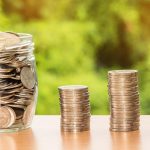 Mão segurando uma cédula de dólar sobre moedas e dinheiro, em uma composição conceitual sobre finanças, depósitos e investimentos.