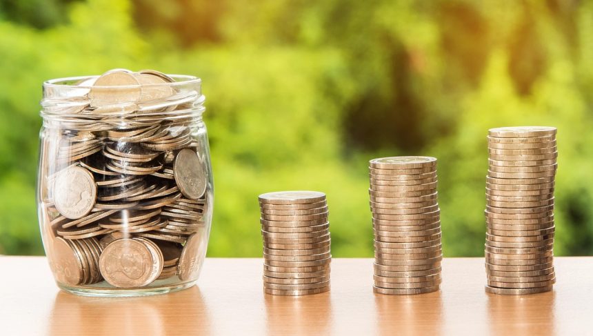 Mão segurando uma cédula de dólar sobre moedas e dinheiro, em uma composição conceitual sobre finanças, depósitos e investimentos.